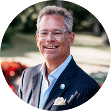 Andrew Goldner in a suit with glasses stands outdoors in a park setting. The background features blurred greenery and vibrant red flowers, evoking a cheerful tone.
