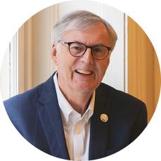 Bob Fealy with glasses and a blue suit he stands near a large window, conveying warmth and friendliness.