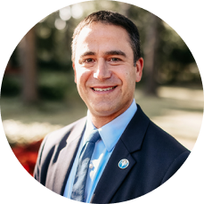 Dr. Brian L. Edlow in a suit and tie smiles warmly outdoors. Sunlit trees and shrubs create a soft, inviting background, suggesting a pleasant, professional setting.
