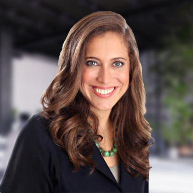 Caitlin Carroll with wavy brown hair wearing a black top and green necklace. Background is blurred with soft, indoor lighting and greenery.