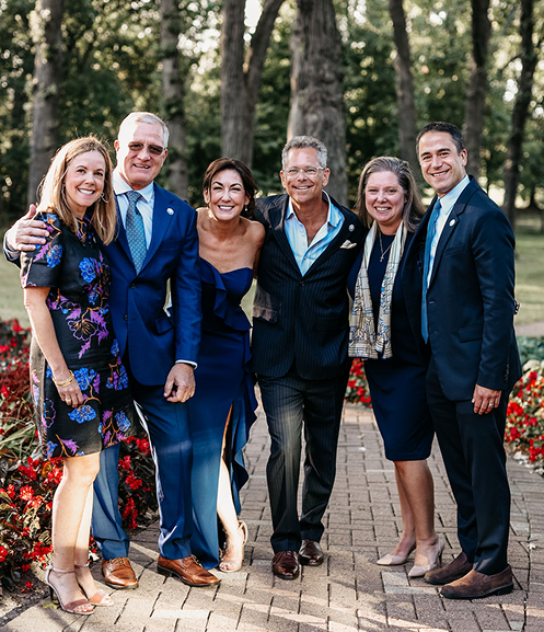 Some of the Invisible Wounds team dressed formally, smiling warmly in a park setting. Trees and vibrant red flowers create a lively and elegant backdrop.