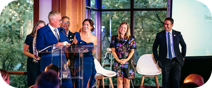 Vice Admiral Tim Szymanski in a blue suit speaks at a podium, surrounded by members of the Invisible Wounds team dressed formally. The setting is a bright, modern venue with large windows.