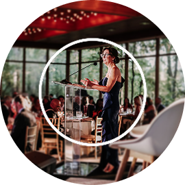 Shannon Connell in an elegant navy dress speaks at a podium at Evening of Honor. Attendees sit at round tables, and large windows reveal a lush green view.