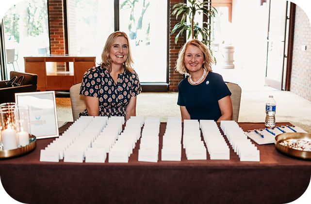 Two women sit at a registration table covered with name cards, clipboards, and a water bottle. They smile warmly in a bright, welcoming room.