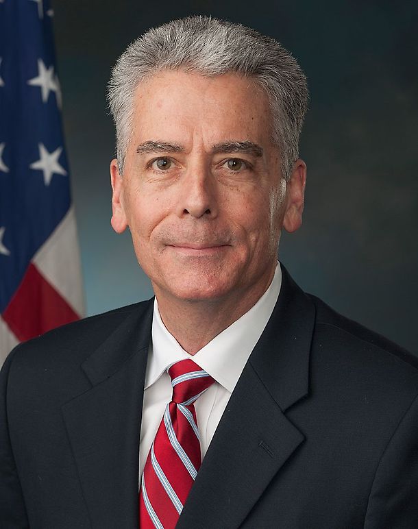 Frank Larkin with short gray hair wearing a dark suit, white shirt, and red striped tie. An American flag is visible in the background. Neutral expression.