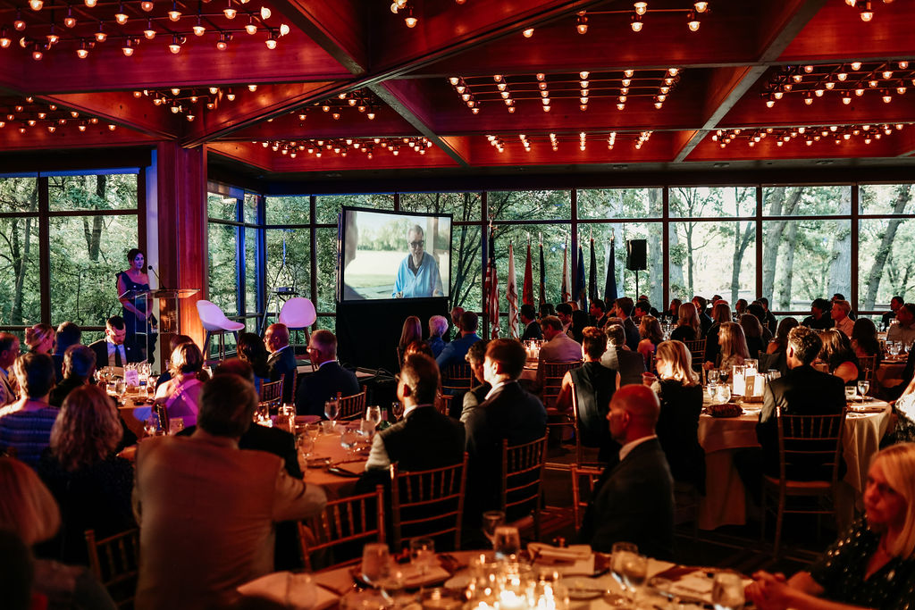 Elegant banquet in a warmly lit room with large windows overlooking trees. Attendees watch a speaker at a podium and a video screen displaying a man.
