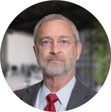 Dr Kelly with glasses and a trimmed beard, wearing a gray suit and red tie, stands confidently in a modern, blurred office setting.